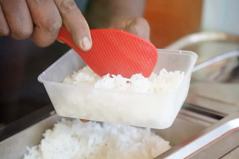 Une main remplit une barquette de riz blanc au-dessus des bacs